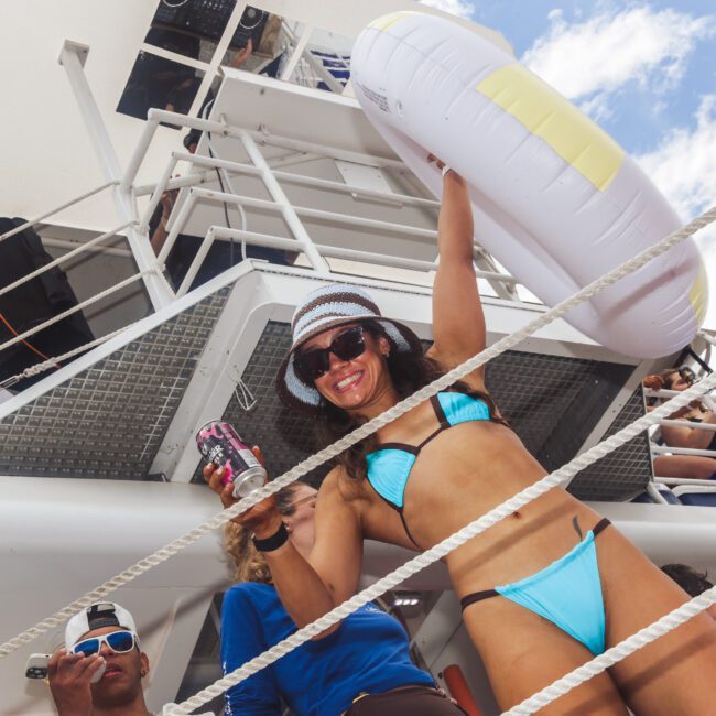 A woman in a blue bikini and sunhat smiles, holding a can and an inflatable ring on a boat deck, with people and blue sky in the background.