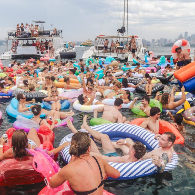 A lively crowd of people floats on colorful inflatable rafts and pool toys in the water, surrounded by boats, enjoying a sunny day party with a city skyline visible in the background.