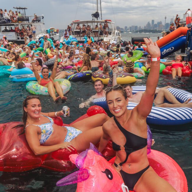 A large group of people relax on colorful inflatable pool floats in the water near boats, smiling and waving at the camera. The scene is lively and festive, with city buildings visible in the background.