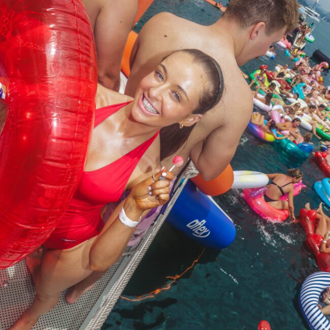 A smiling woman in a red swimsuit holds a red inflatable ring and a lollipop on a dock, surrounded by people and colorful inflatables at a lively pool party on the water.