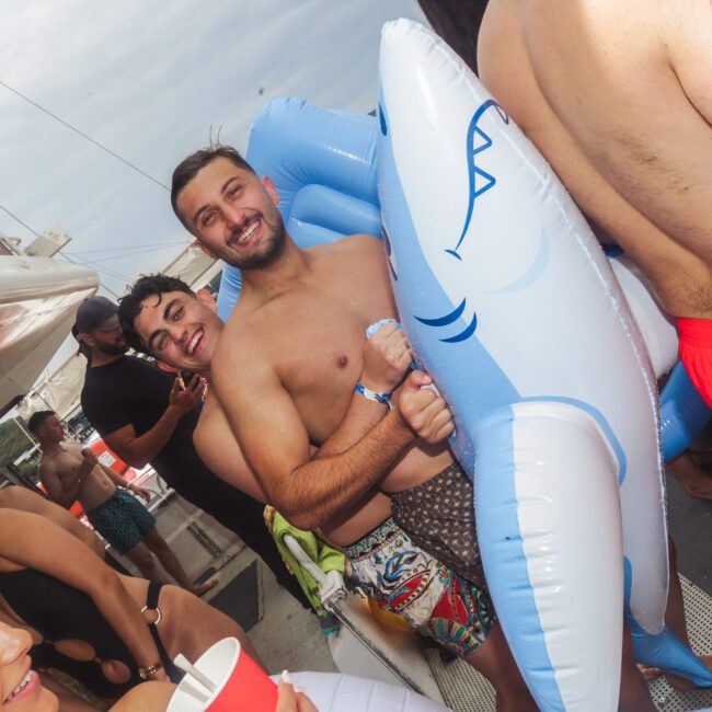 A group of people in swimwear enjoy a party on a boat. One man smiles at the camera while holding a large inflatable shark. Others are chatting, laughing, and holding drinks, with a lively and festive atmosphere.