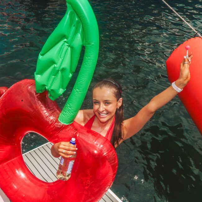 A smiling woman on a boat holds a drink and a lollipop while wearing a red cherry-shaped pool float. Other people are swimming and lounging on floats in the water nearby. The scene is lively and summery.