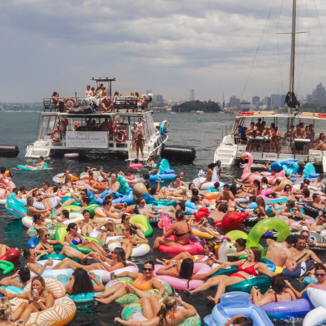 A large crowd of people on colorful inflatable floats relax and socialize on the water, with two boats nearby, under a cloudy sky with a city skyline in the background.