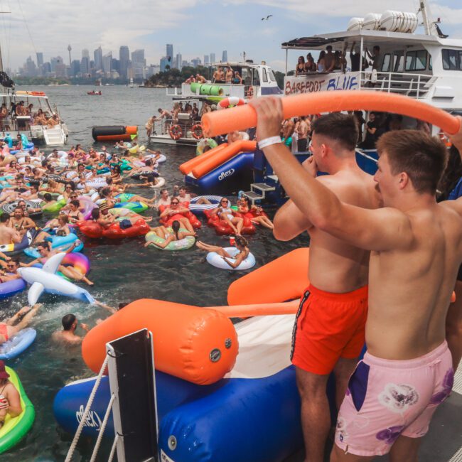 Two men in swimwear stand on a boat holding pool noodles, overlooking a crowded water party with many people on colorful inflatables. Boats and a city skyline are visible in the background.