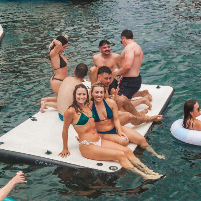 A group of people in swimwear relax on a floating platform and inflatable ring in a body of water. Some are sitting and talking, while others are lounging and smiling at the camera, enjoying a sunny day.