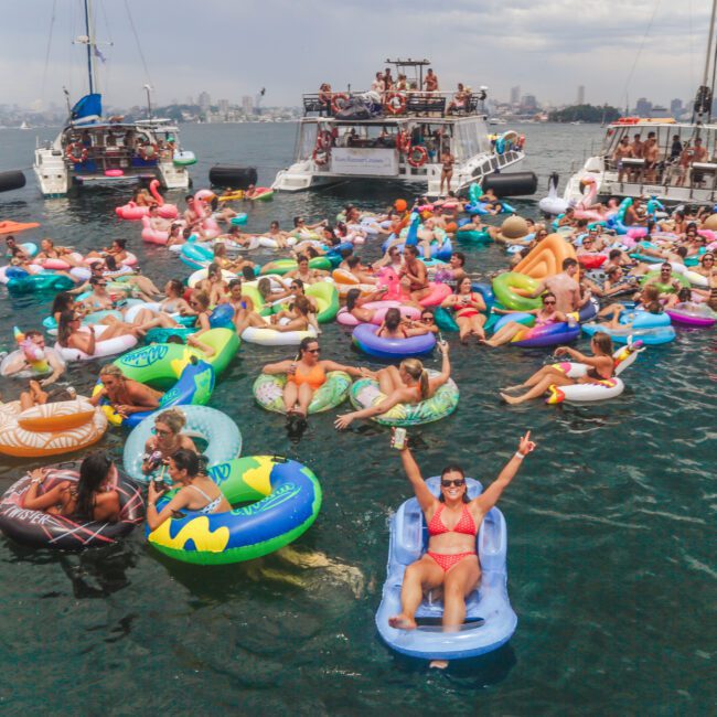 A large crowd of people on colorful inflatable floaties relax and socialize in the water near several anchored party boats on a sunny day, with a city skyline visible in the background.