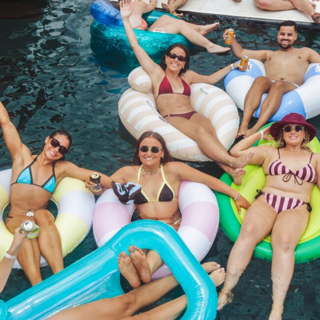 Five people in swimsuits relax on colorful pool floats in the water, smiling and raising their arms, each holding drinks. The scene is lively and joyful, suggesting a fun summer outing with friends.