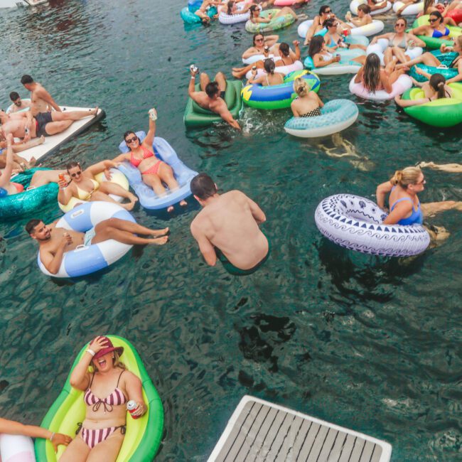 A group of people relax in colorful pool floats and swim in the water at a lively outdoor party. One person is mid-air, jumping into the water, while others chat, sunbathe, and enjoy the atmosphere.