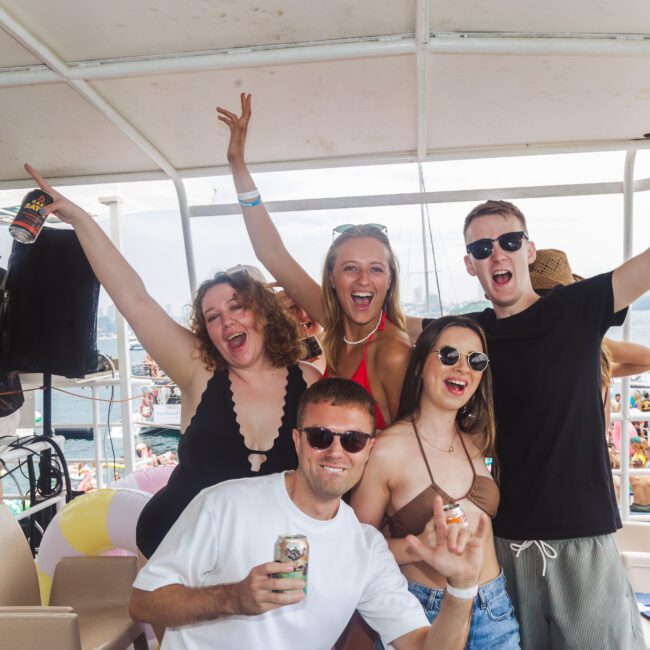 Five young adults in swimsuits and sunglasses pose, smiling and raising their arms, holding drinks on a crowded boat party with people and city skyline in the background.