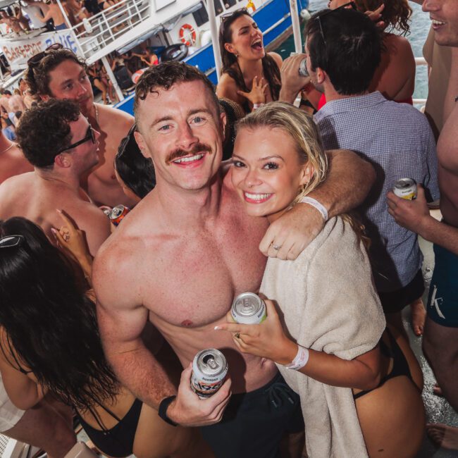 A group of people in swimsuits smile and hold drinks during a lively boat party, with two people in the foreground hugging and posing for the camera. A large boat and water are visible in the background.