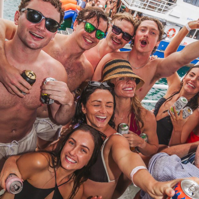 A group of young adults in swimwear smile and cheer with drinks in hand at a lively boat party on the water, surrounded by other people, boats, and colorful pool floats.
