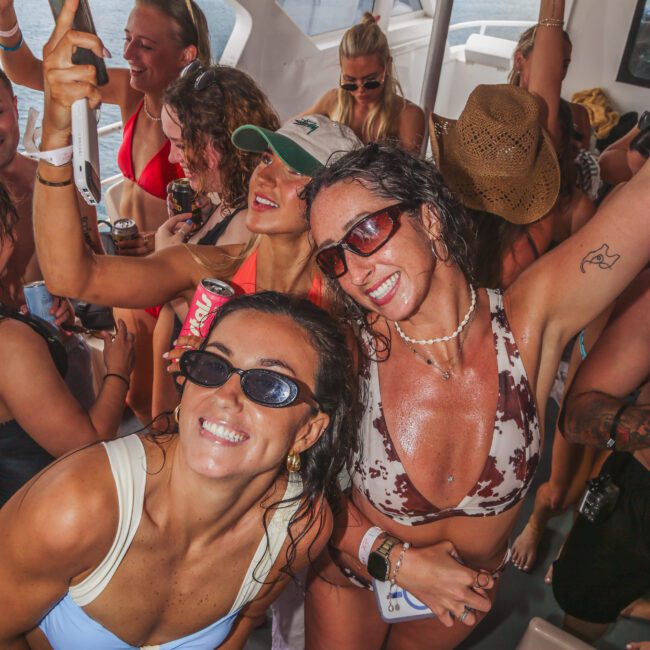 A group of women in swimsuits and sunglasses dance and smile on a boat, taking selfies and enjoying drinks. Sunlight streams in, and the atmosphere is lively and festive.