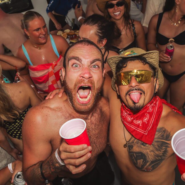 A group of people in swimsuits party indoors. Two men in front, one yelling with a beard and holding a red cup, the other wearing sunglasses, a cowboy hat, and a red bandana, both smiling and excited.
