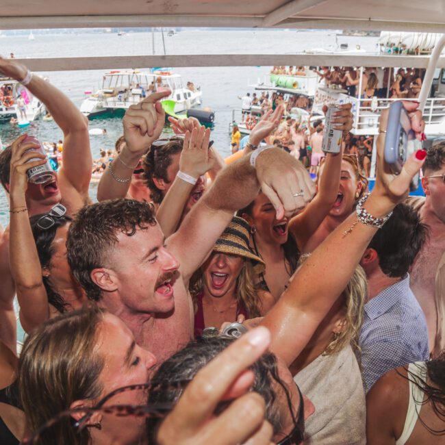 A large group of young adults in swimsuits cheer and raise drinks while partying on a crowded boat, with more people and boats visible on the water in the background. Everyone appears excited and energetic.
