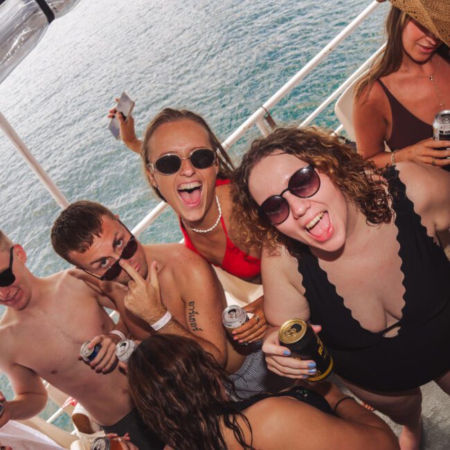 A group of young adults in swimsuits are smiling and posing together on a boat, holding drinks. The ocean is visible in the background, and everyone appears to be enjoying a lively, sunny day.