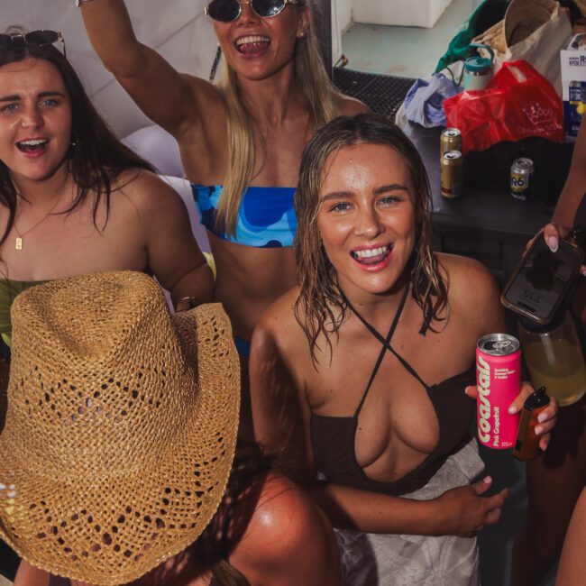 A group of young women in swimsuits and summer attire smiling and enjoying themselves at a party on a boat. One woman holds a drink, and another wears a straw hat. There are drinks and snacks on the table behind them.