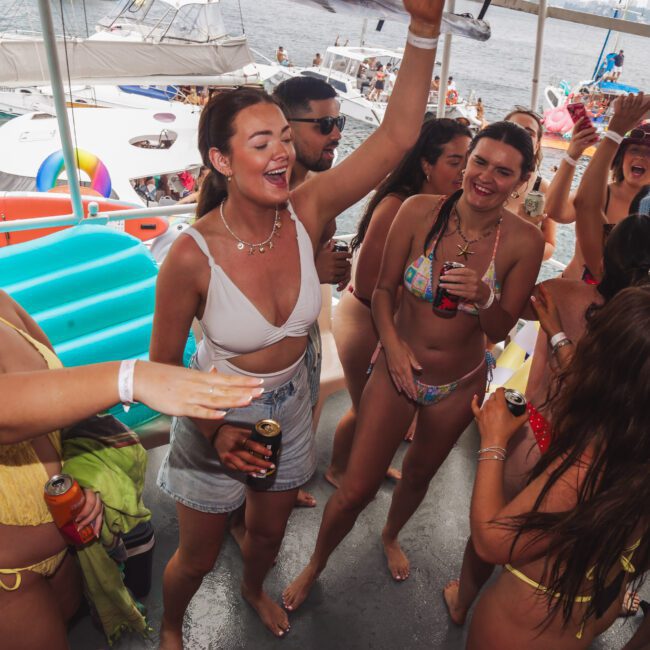 A group of people in swimsuits dance, laugh, and hold drinks on a boat deck under a canopy, with water, yachts, and a city skyline visible in the background. It appears to be a lively summer party.