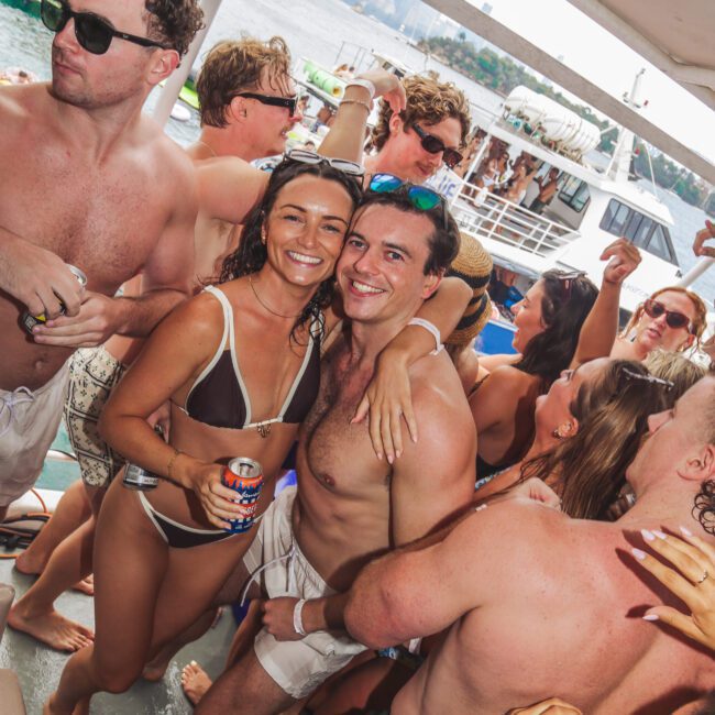 A group of young adults in swimsuits are smiling and dancing on a crowded boat, holding drinks. The background shows water, other boats, and a city skyline under a bright, sunny sky.