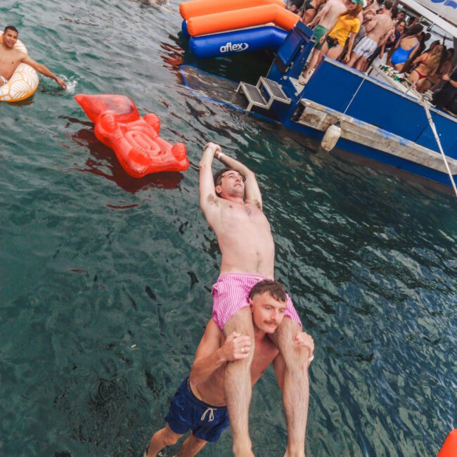 Two men playfully jump off a dock into the water; one is holding the other overhead. People on inflatables and a crowded party boat are in the background, with a city skyline visible through the haze.