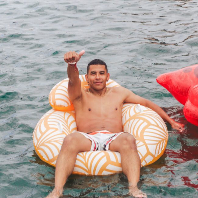 A man in swim trunks sits on an orange and white float in the water, smiling and giving a thumbs up. Another red float is partially visible beside him.