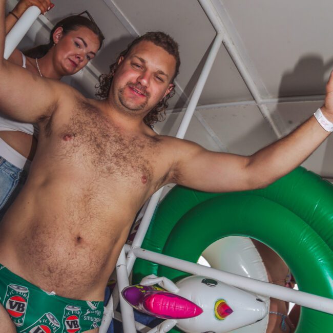 A man wearing VB beer-branded swim briefs poses with his arms outstretched, standing near inflatable pool rings and a unicorn float. A woman with sunglasses on her head stands behind him, both on what appears to be a boat or pool deck.