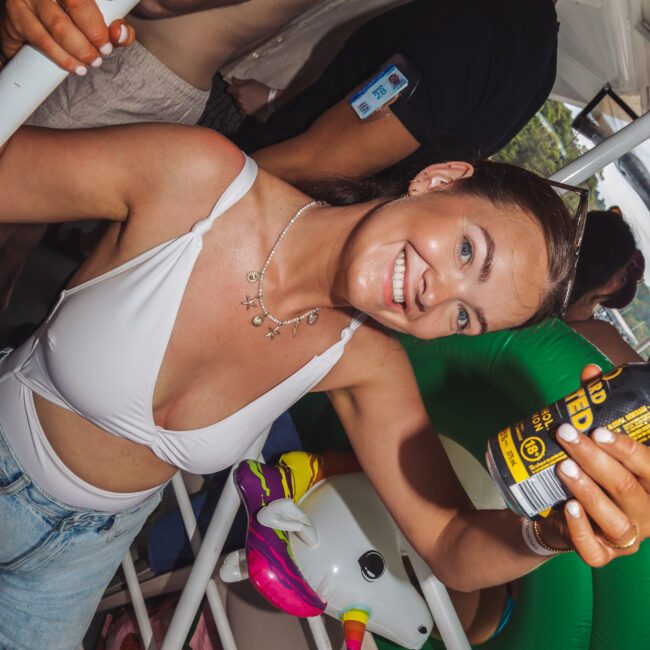 A smiling woman in a white bikini top and denim shorts holds up a can at a crowded pool or boat party. People and inflatable pool toys, including a unicorn float, are in the background.