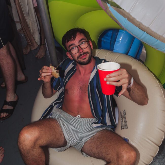 A man with glasses sits in an inflatable pool seat at a party, holding a sandwich and a red cup. He wears a striped shirt, shorts, and appears relaxed. Inflatable pool toys and other people are visible around him.