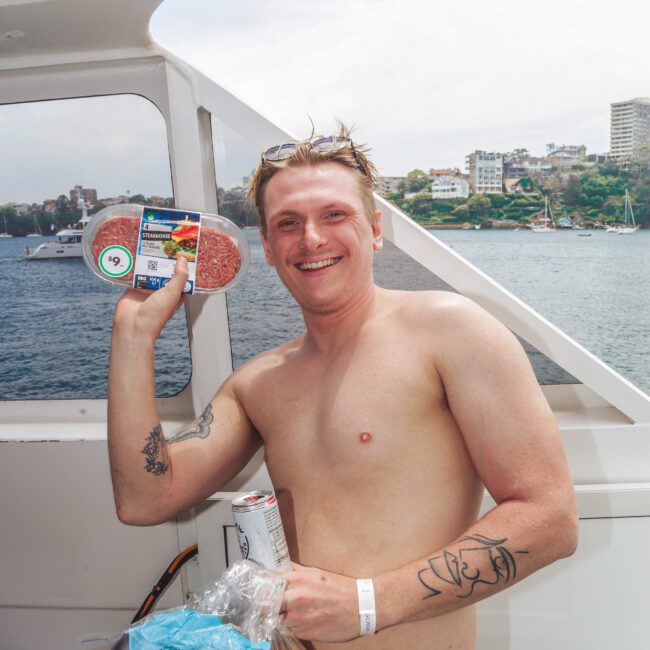 A smiling shirtless man with tattoos holds up a package of hamburger patties and hot dog buns on a boat, with city buildings and water visible in the background.