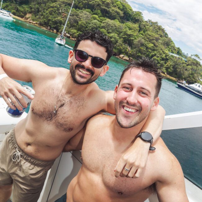 Two smiling men in swim trunks stand close together on a boat, with one holding a drink. Behind them, there is a scenic view of water, trees, and anchored boats under a partly cloudy sky.