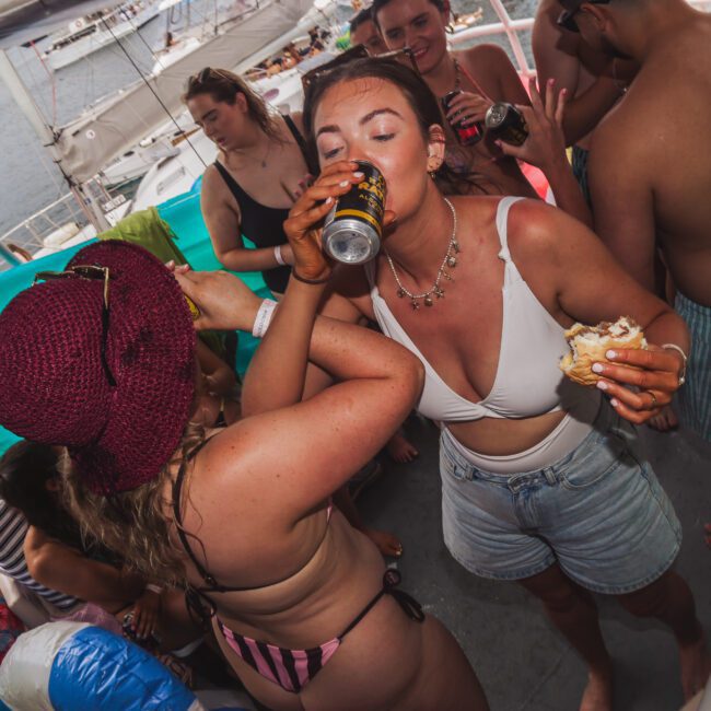A group of people in swimsuits enjoy drinks and food on a boat. One woman in a red hat drinks from a can, while another woman in shorts and a necklace eats a sandwich and drinks. Boats are visible on the water in the background.