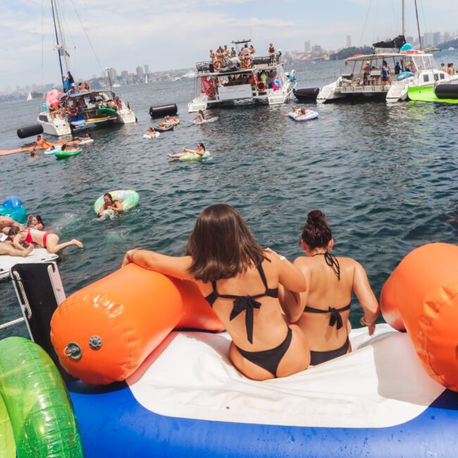 Two women in black bikinis sit on a large inflatable float in the water, facing a group of boats and people on floats. The scene is lively with many enjoying a sunny day on the water.