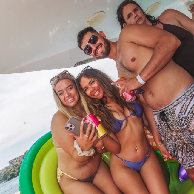 Four people in swimwear smile and pose together on a boat with drinks in hand. Two women sit on an inflatable float, while two men stand behind them. Bright daylight and water are visible in the background.