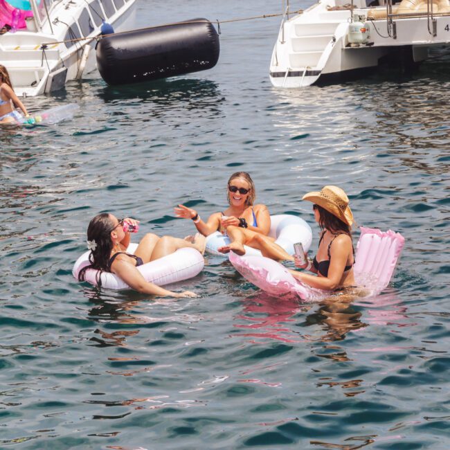Three women relax on inflatable floats in the water near boats, laughing and chatting under sunny skies. Other people lounge on the boats or in the water, enjoying a lively, fun day.
