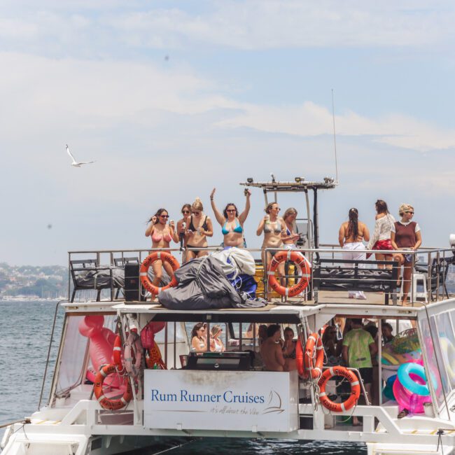 A group of people in swimwear are dancing and enjoying themselves on the upper deck of a Rum Runner Cruises boat on the water, with life rings attached to the boat and city buildings visible in the background.