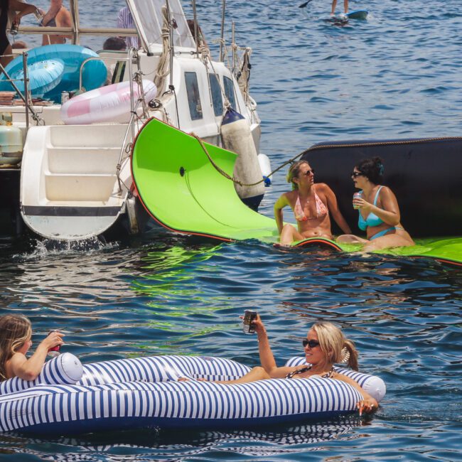 Several people relax on floating mats and inflatable rafts near a boat on the water. Two women are chatting on a green mat, while two others lounge on a striped inflatable, holding drinks and enjoying the sun.