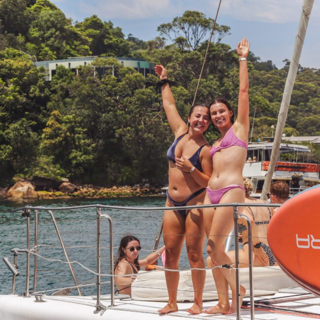 Two women in bikinis smiling and raising their arms stand on a boat deck, with trees and buildings visible in the background. A red paddleboard is attached to the railing, and other people are relaxing nearby.