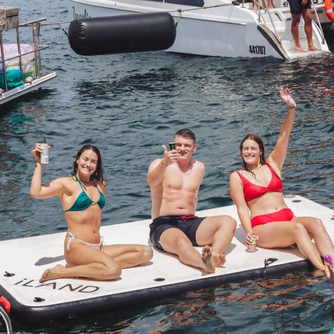 Three people in swimsuits sit on a floating mat in the water, smiling and waving while holding drinks. Boats and inflatables surround them, creating a lively and cheerful summer scene.