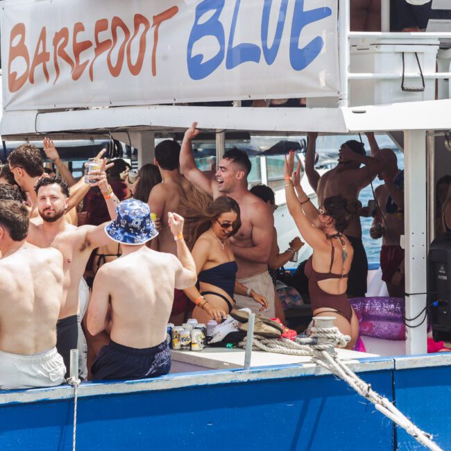 A group of young adults in swimsuits dance and celebrate on a crowded party boat called "Barefoot Blue," under sunny weather, with drinks in hand and a festive atmosphere.
