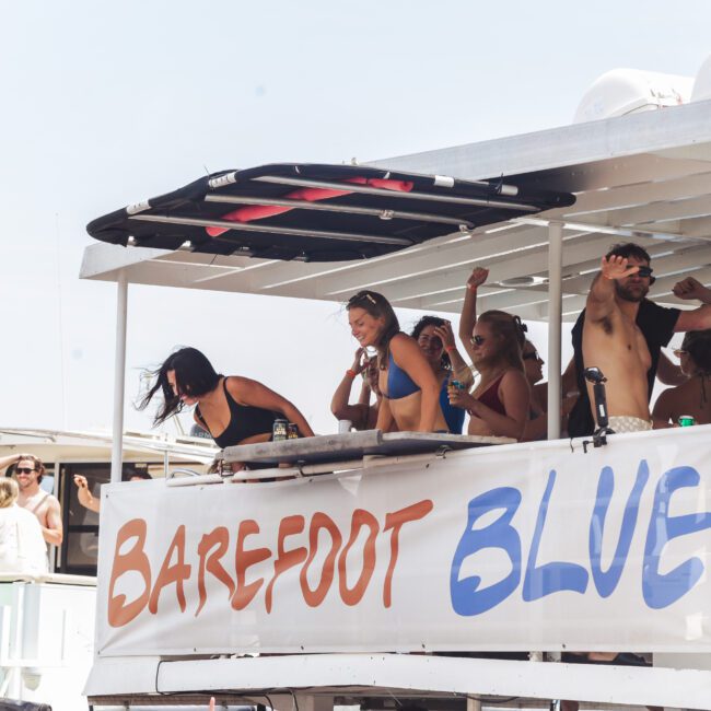 A group of people in swimwear dance and socialize on a boat named "Barefoot Blue" during a sunny day, with other boats and people visible in the background.