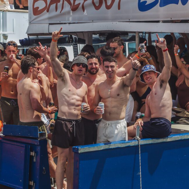 A group of young people in swimwear enjoy a lively boat party under the sun, with some holding drinks and smiling at the camera, while others socialize in the background.