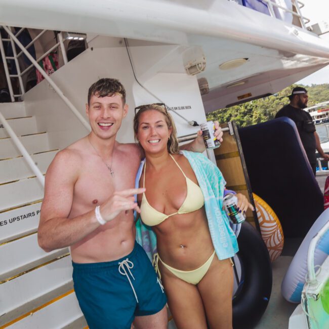 A man and woman in swimwear smile and pose together on a boat deck, surrounded by inflatables and other people. Stairs with a "WET BODIES UPSTAIRS PLEASE" sign are in the background.
