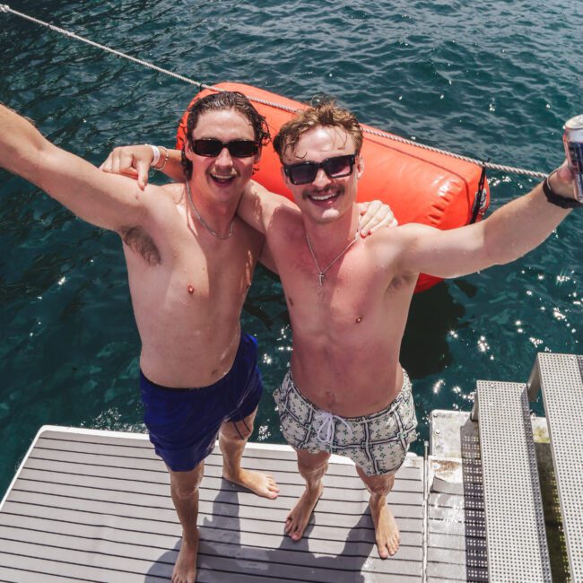 Two smiling men in swim trunks, sunglasses, and hats stand on a dock platform, arms around each other, enjoying a sunny day by the water with a red inflatable raft and blue sea in the background.