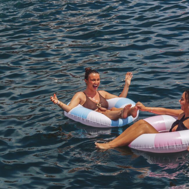 Two women in swimsuits smile and hold hands while relaxing on inflatable pool rings in blue water, enjoying a sunny day.