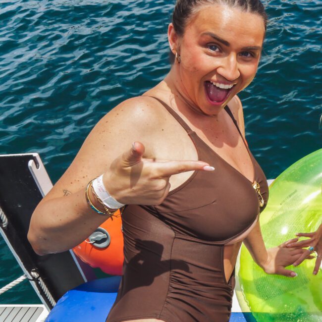 A woman in a brown one-piece swimsuit smiles and points excitedly while standing on a boat next to colorful inflatables, with blue water in the background.
