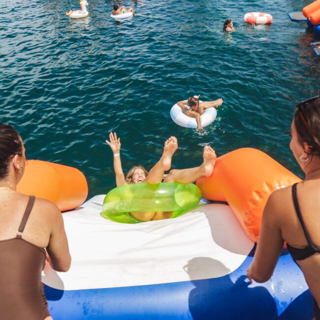 Two women watch as a person joyfully slides down an inflatable water slide into a blue body of water, surrounded by others floating on colorful inflatables.