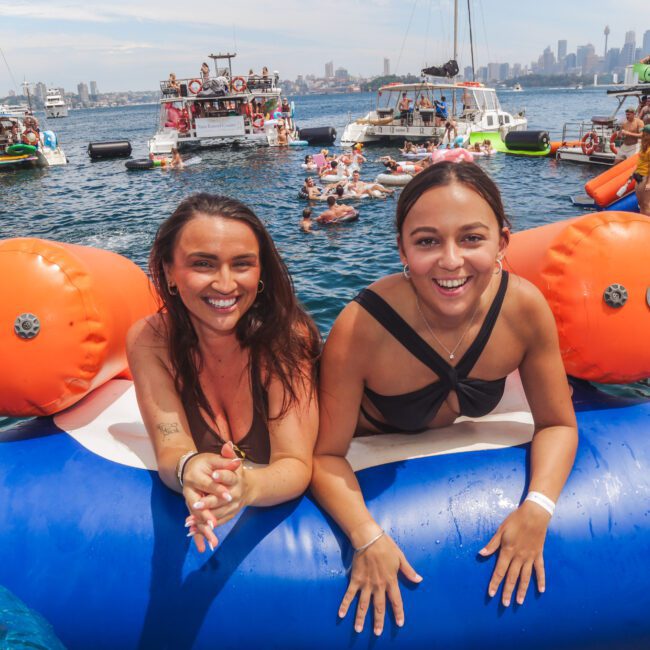 Two women in swimsuits smile and pose on an inflatable float in the water. Boats and people enjoying a sunny day on the water are visible in the background, with a city skyline in the distance.
