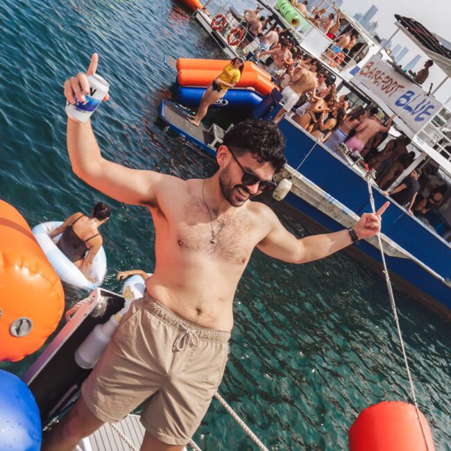 A man in sunglasses and shorts smiles, holding a drink and leaning on a rope, standing on a docked boat with colorful inflatables. Behind him, people party on boats with a city skyline in the background.