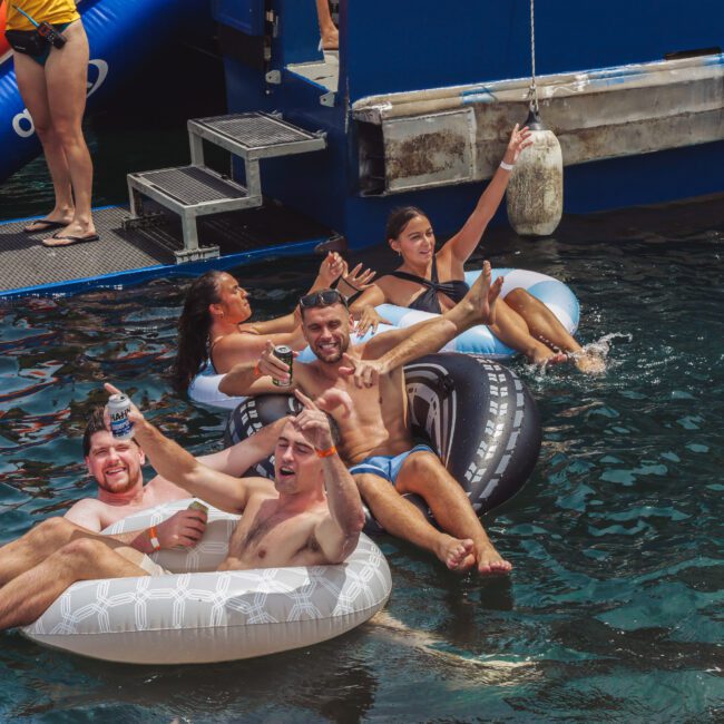 Four people relax on inflatable tubes in the water near a dock, smiling and holding drinks. A woman stands on the dock above them, and the scene appears cheerful and summery.