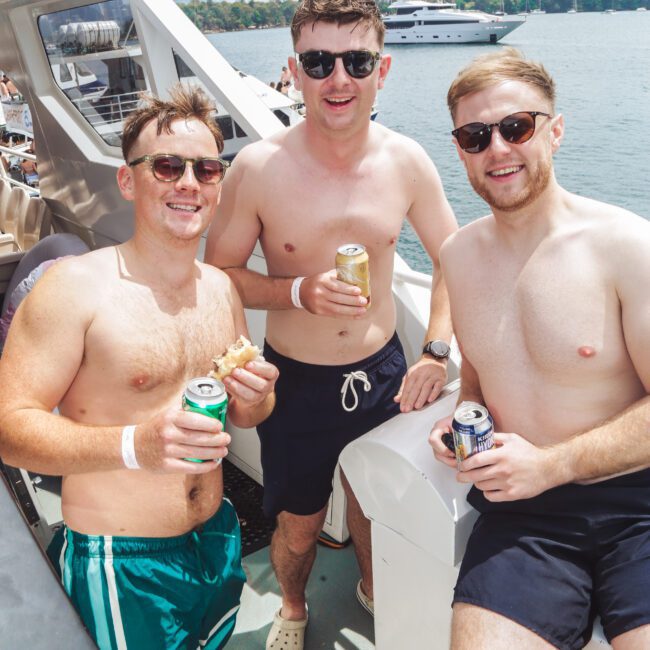 Three young men in sunglasses and swim trunks smile and hold drinks while relaxing on a boat, with water, another boat, and a city skyline visible in the background on a sunny day.