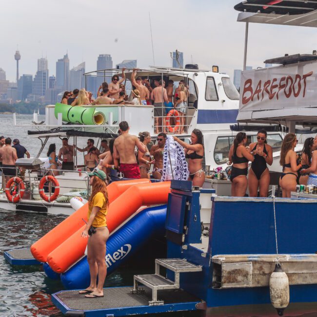 A group of people in swimsuits enjoy a lively boat party with water slides, inflatables, and drinks on a sunny day. Several boats are anchored together on the water with a city skyline in the background.